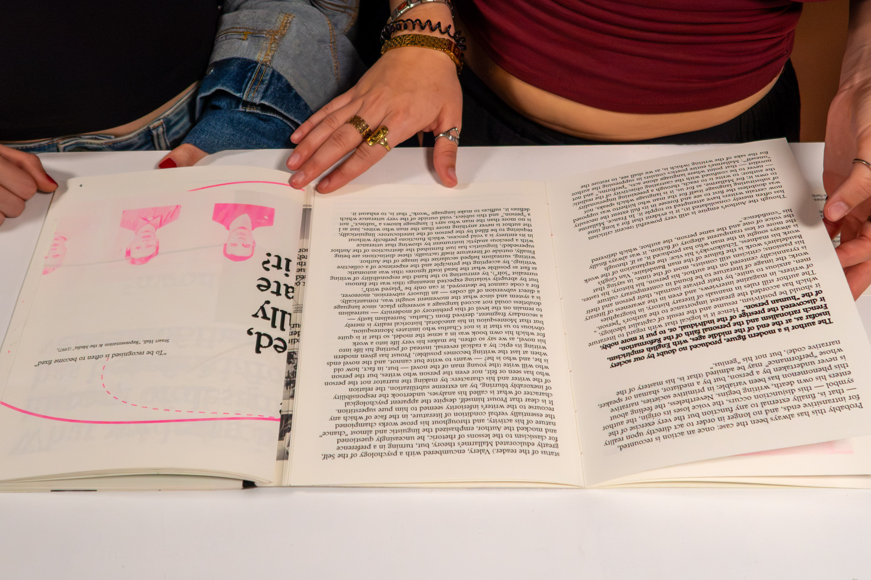 Two people reading an open risograph-printed publication on a table, pink and black text visible across both pages.