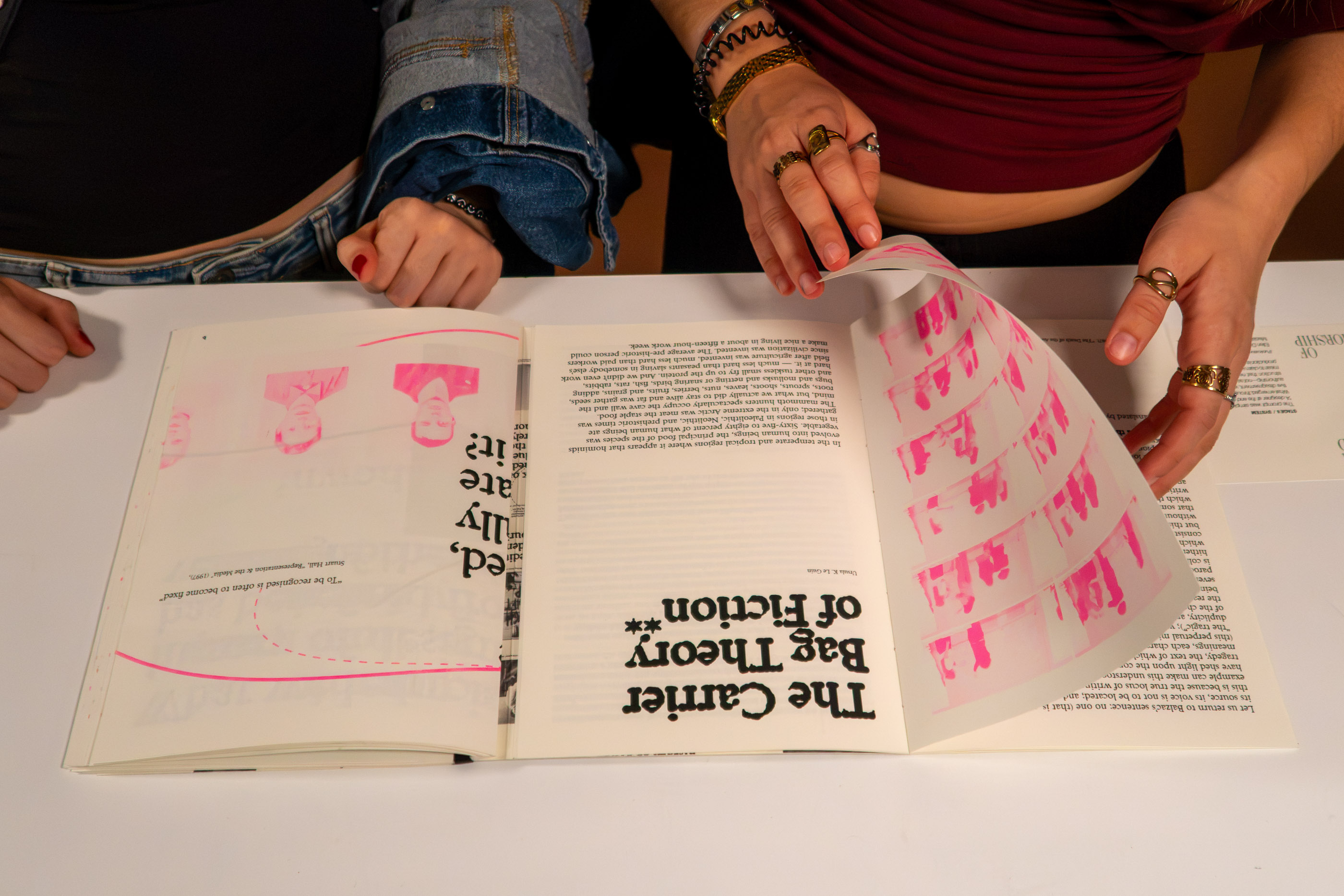 Hands opening the publication on a table, spine visible, pink risograph illustrations on the left page.