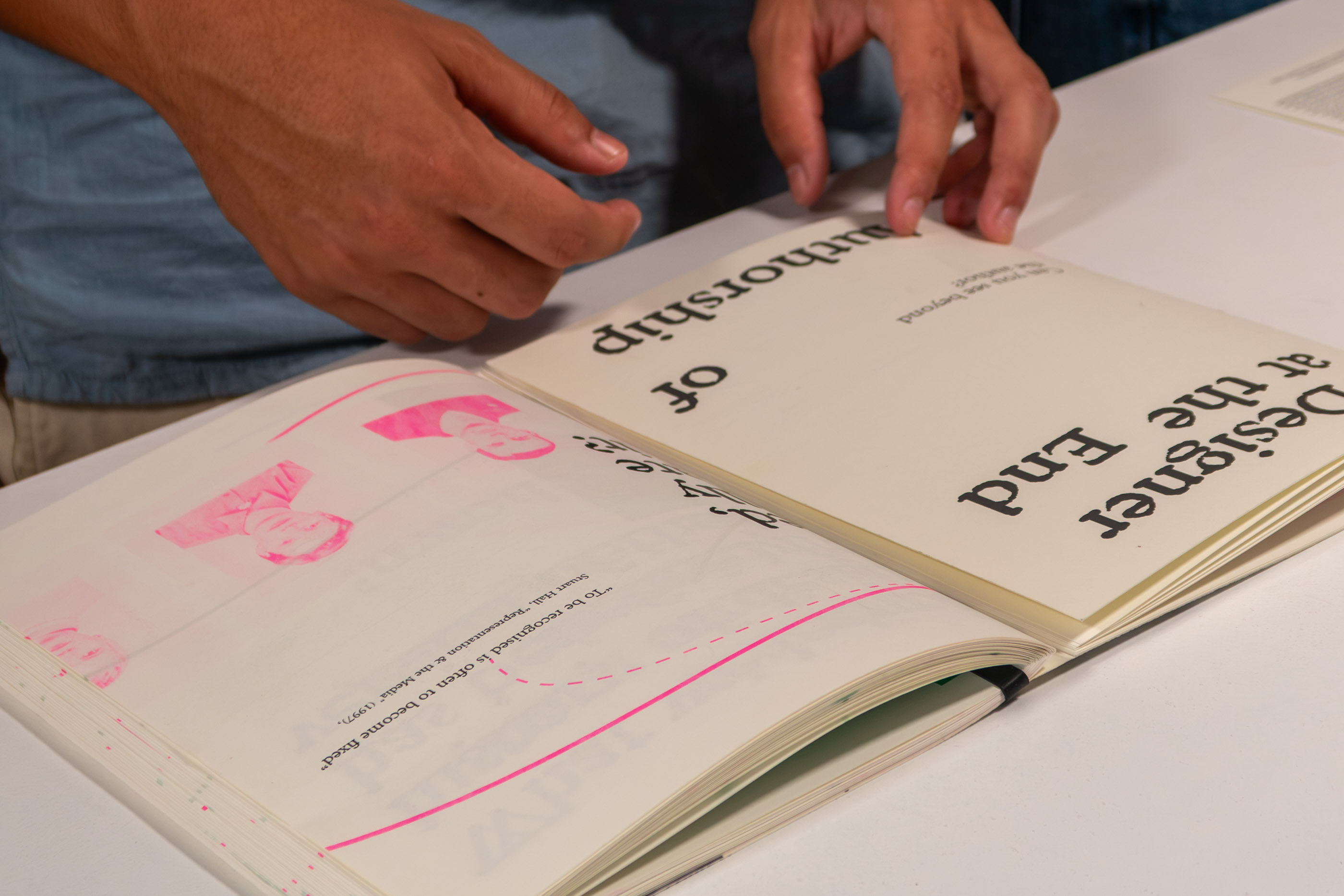 Two people at a table turning a page, revealing pink risograph print on the reverse.